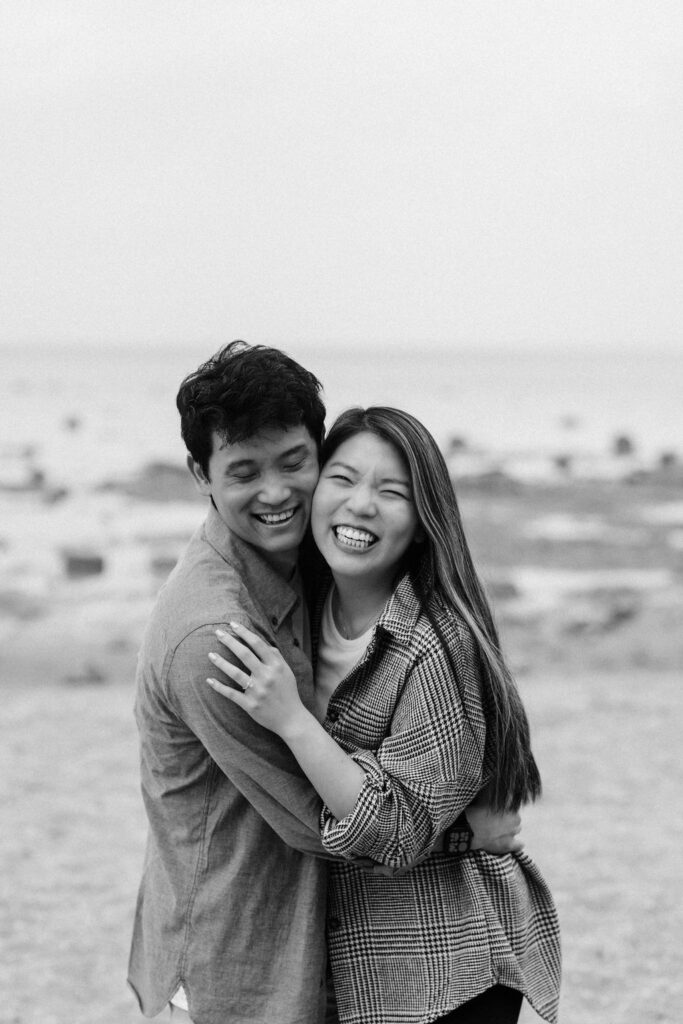 Black and white candid photo of a couple smiling and laughing at Old Mission Lighthouse on the beach.