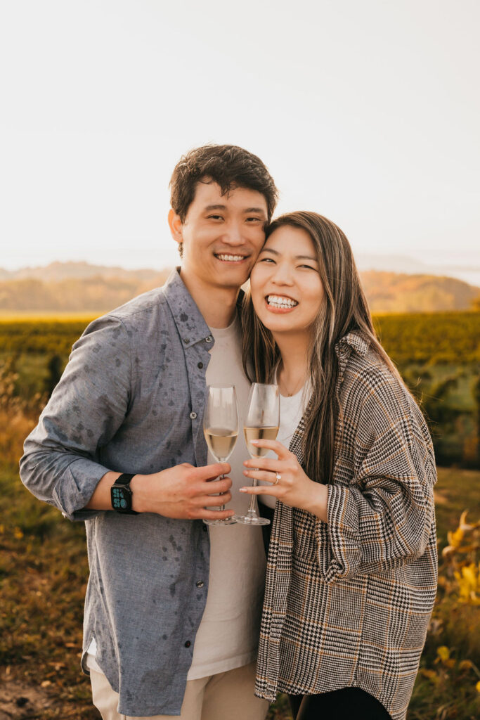 Couple smiling and holding champagne glasses during their Traverse City Michigan proposal photos at The Overlook.
