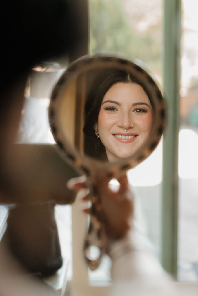 bride looking in a handheld mirror smiling while getting ready for her wedding day